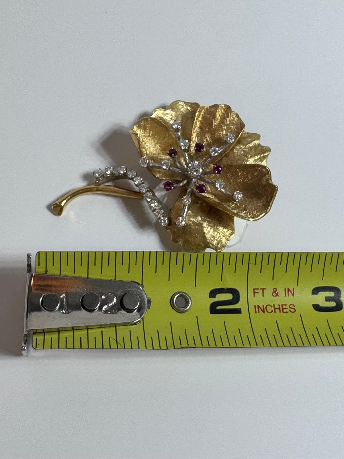 Gold flower-shaped brooch with gemstones next to a measuring tape on a white background
