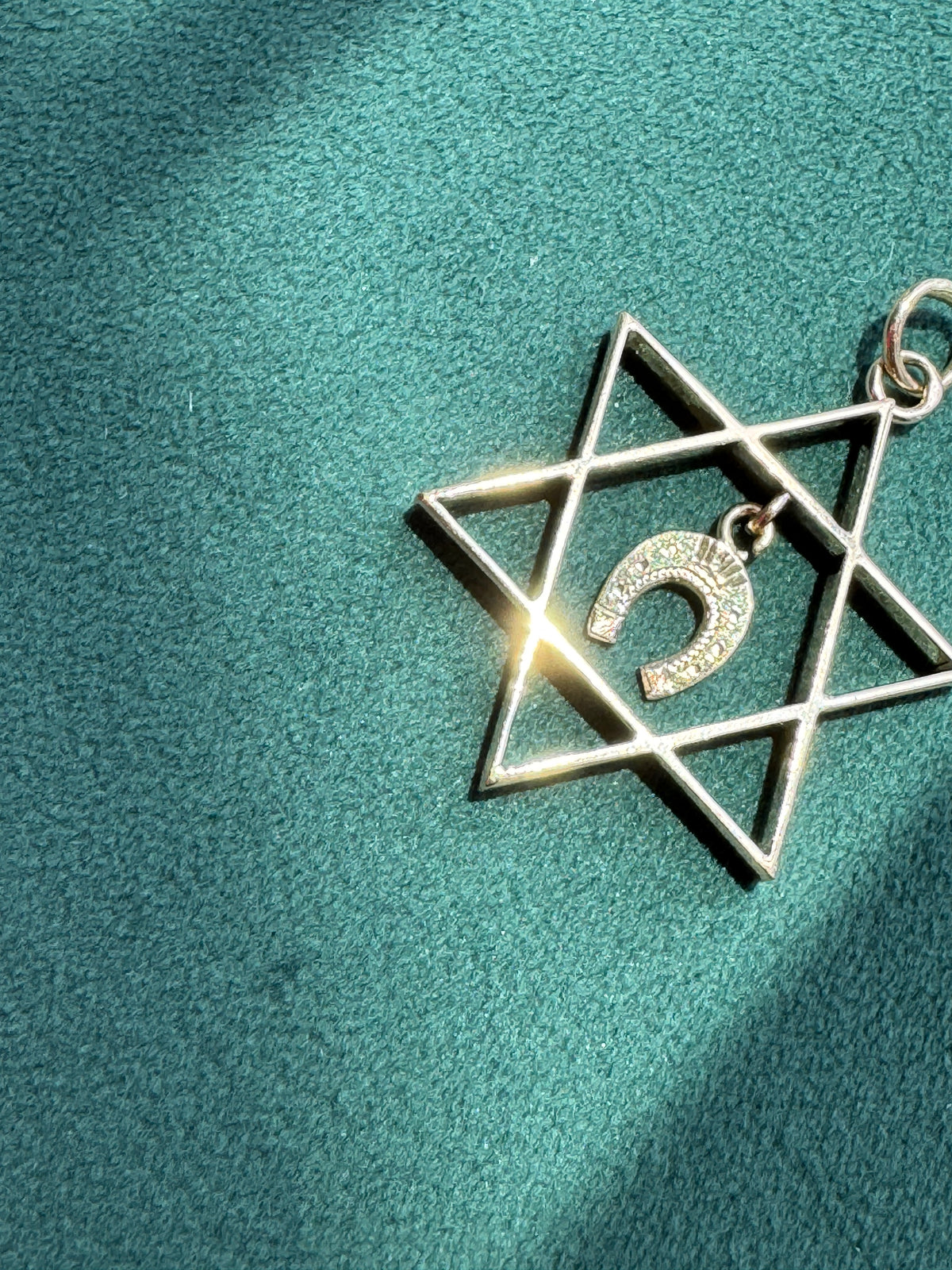 A silver-toned Star of David pendant with a small Shema letter charm, displayed on a textured teal fabric background.