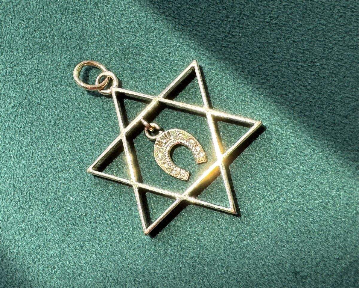 A gold-toned Star of David pendant with a small Hebrew letter charm in the center, resting on a textured green surface with soft shadows.