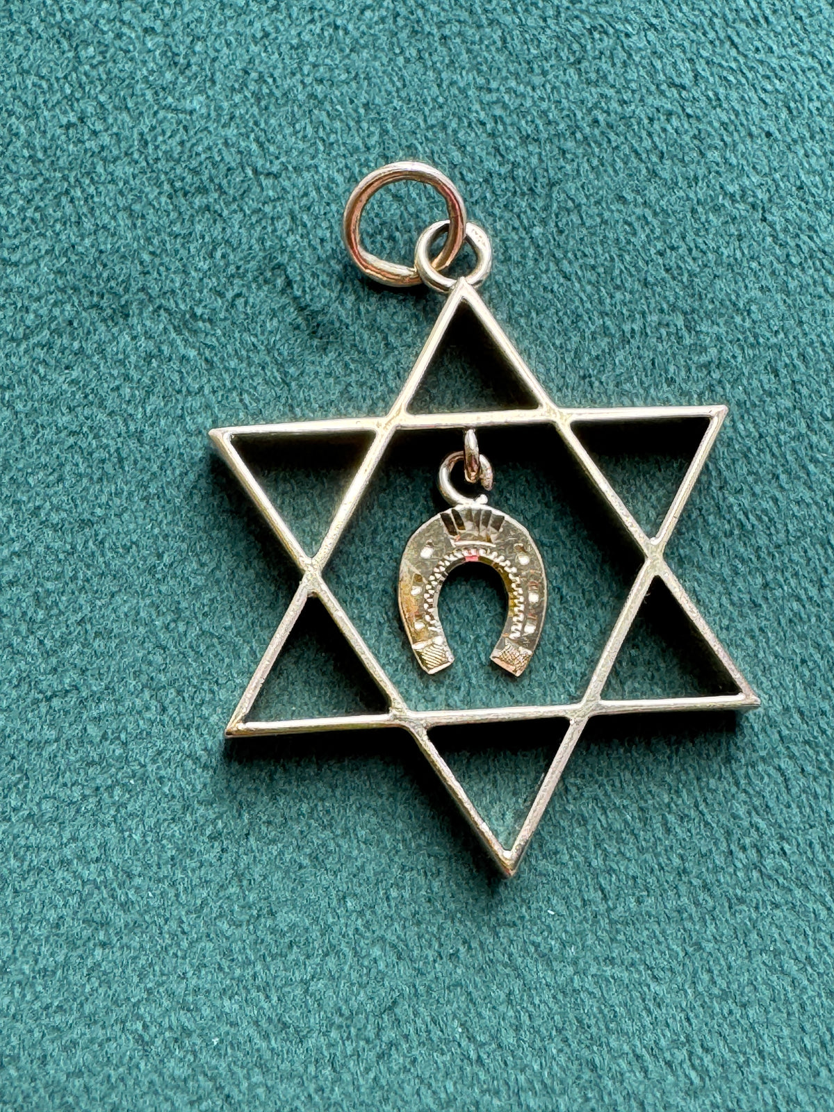 A silver-tone Star of David pendant featuring a small hanging horseshoe charm with a crown detail, displayed on a textured teal background.