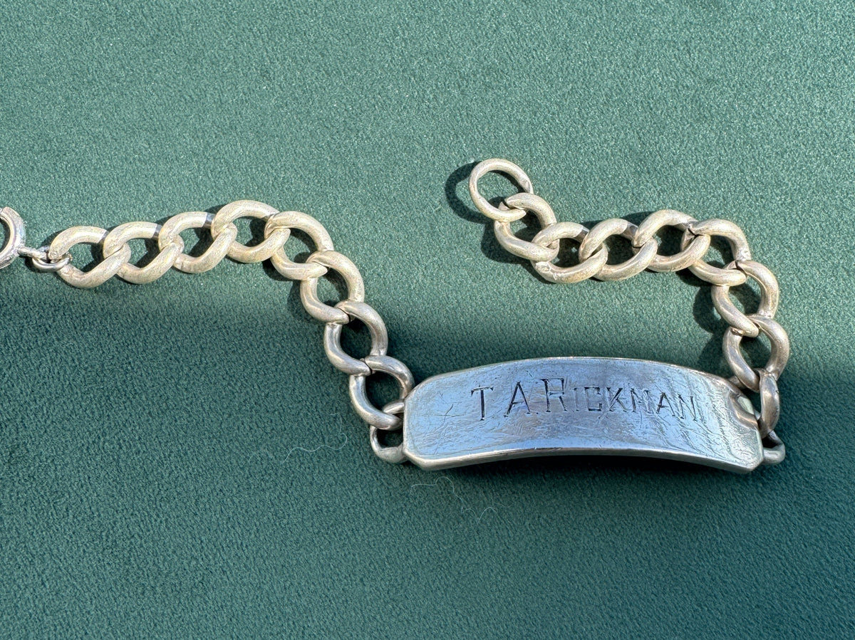 A silver nameplate bracelet with a textured, engraved plate connected to a curb chain, displayed on a dark green textured background.