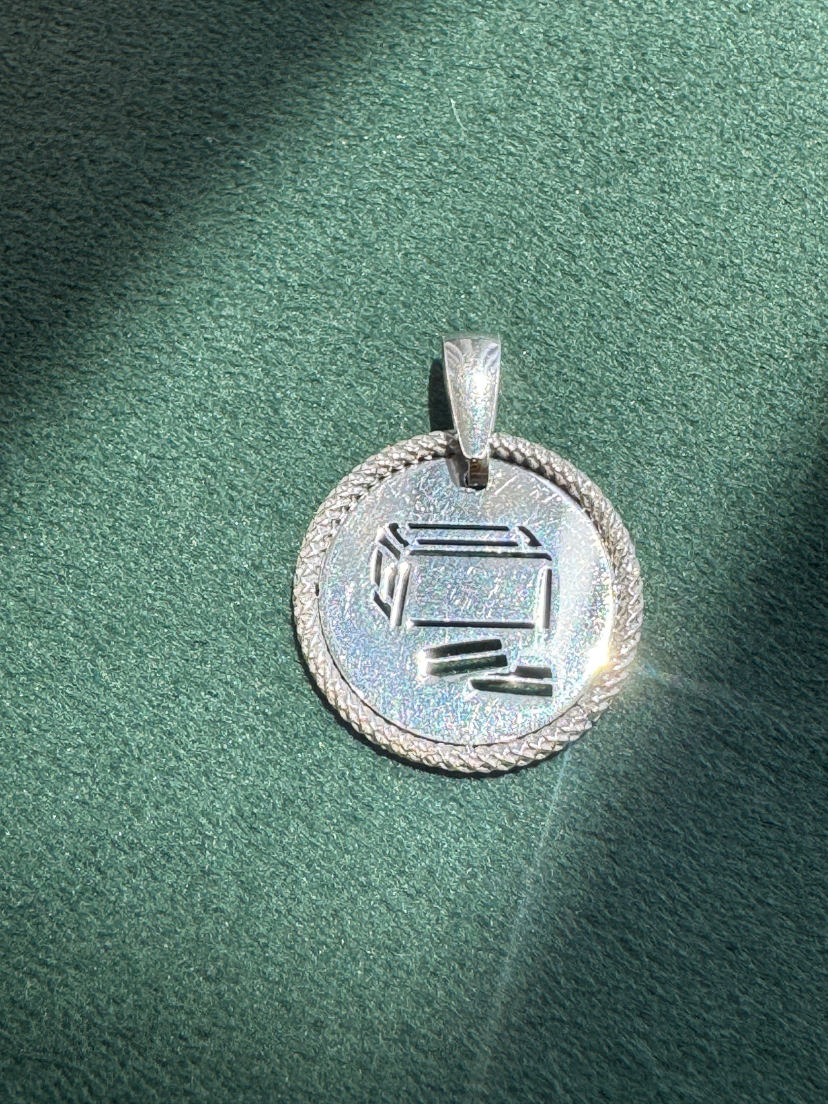 A silver pendant with a circular design, featuring a Hebrew letter 'Shin' engraved in the center and a braided border, resting on a textured green surface.