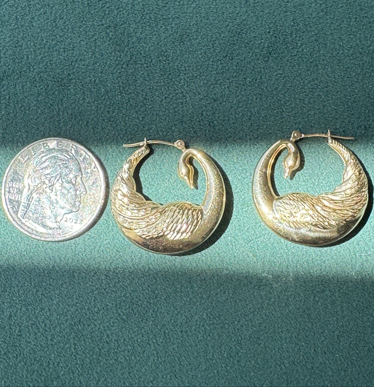 A pair of gold crescent-shaped hoop earrings with a textured, feather-like design, displayed next to a US quarter coin for scale on a dark green textured background.