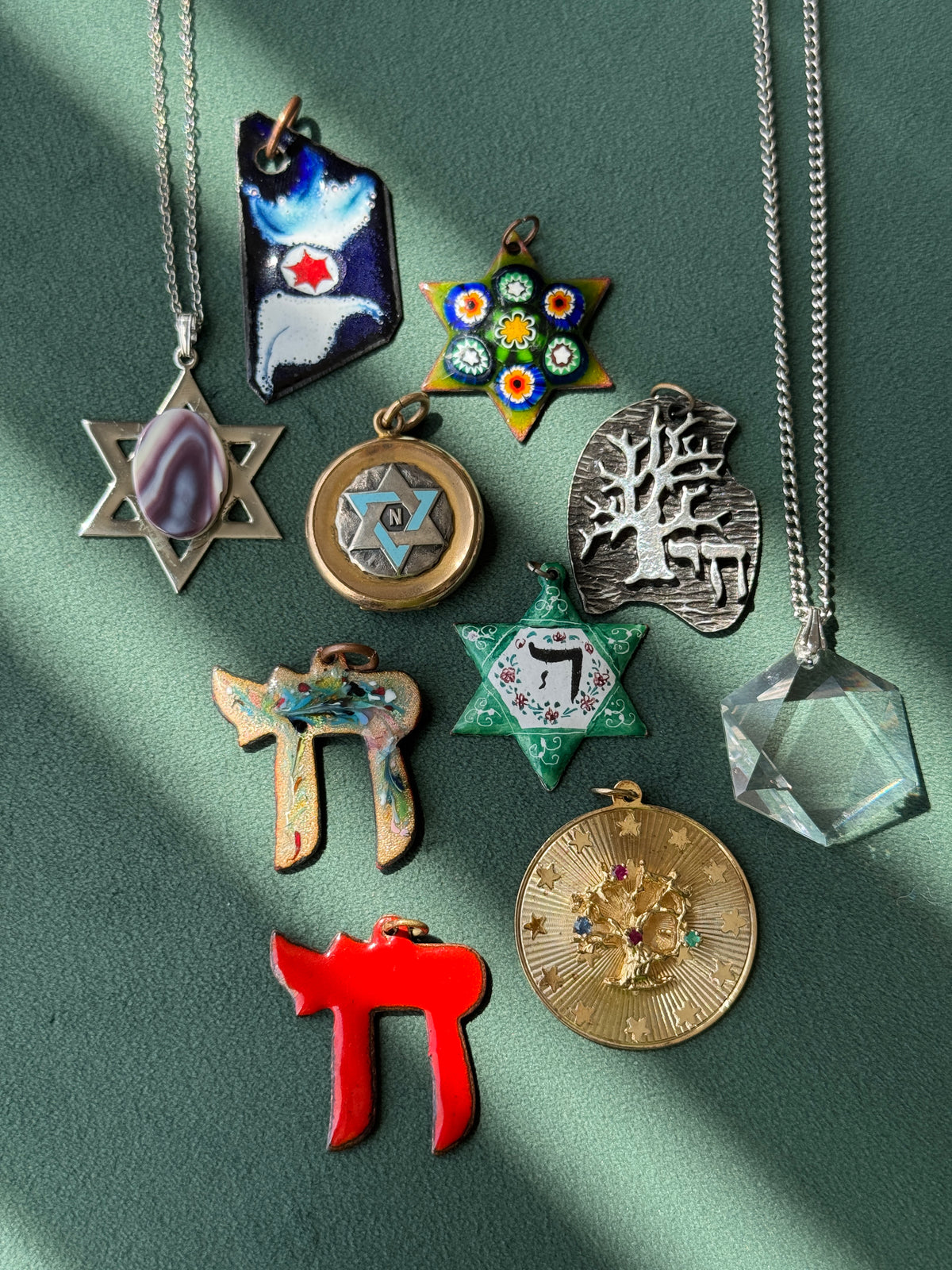 A collection of nine colorful Judaica-themed pendants on silver chains, arranged on a textured green background with diagonal shadows. The pendants include a Star of David with a purple stone, a blue and white Hebrew letter pendant, a colorful Star of David, a silver Tree of Life pendant, a gold Star of David with a Hebrew letter, a Hebrew letter pendant, a red Hebrew letter pendant, a gold medallion pendant, and a clear faceted crystal pendant.
